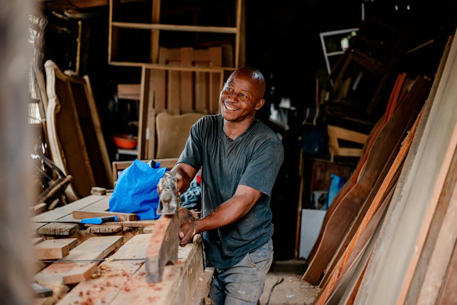 Handwerker hobelt lachend ein Brett in seiner Werkstatt.