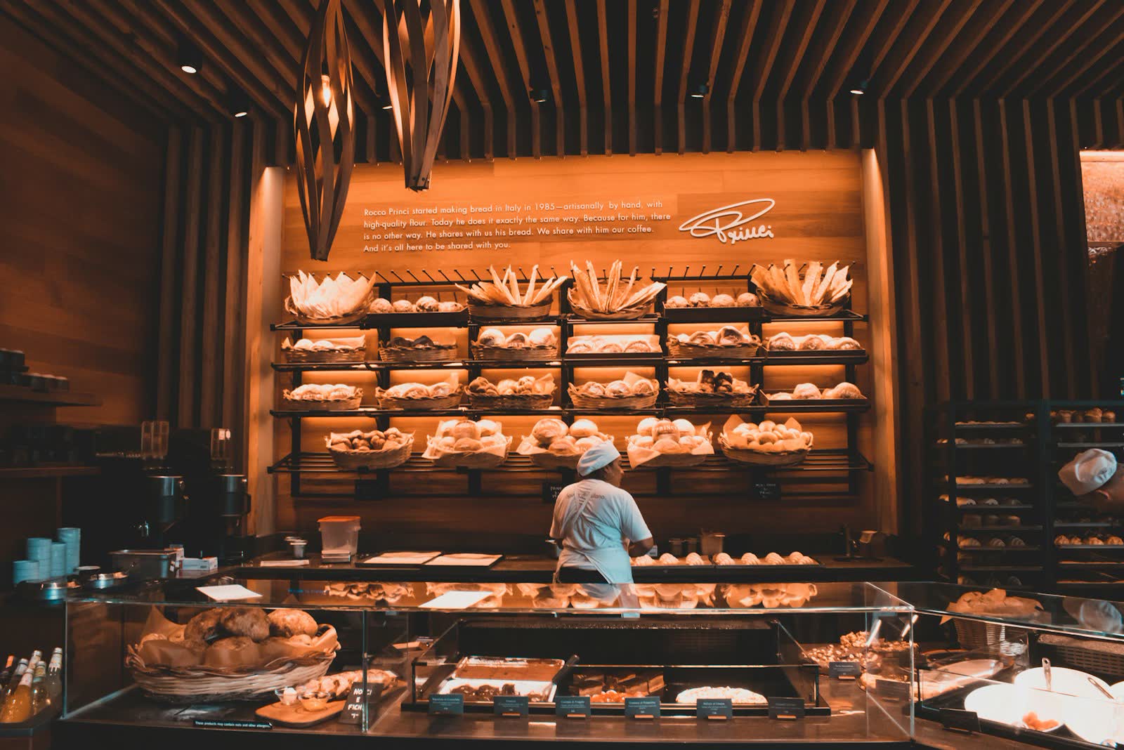 Warm beleuchtete Bäckerei mit vollen Regalen und einem Bäcker hinter der Theke.